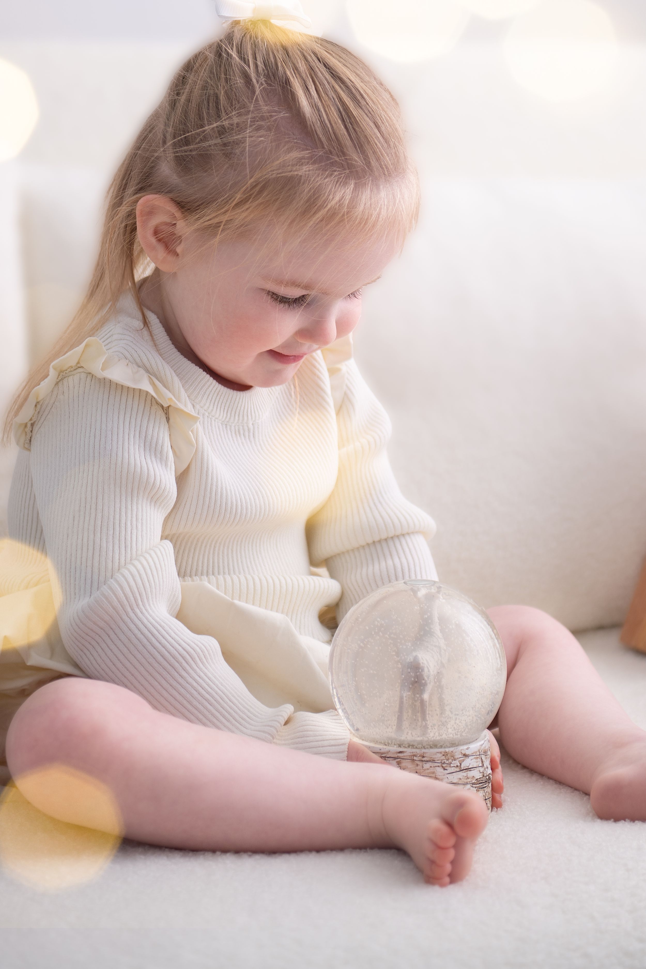 A child in a white sweater lies on a soft white carpet while playing with a toy bunny in a bright room.