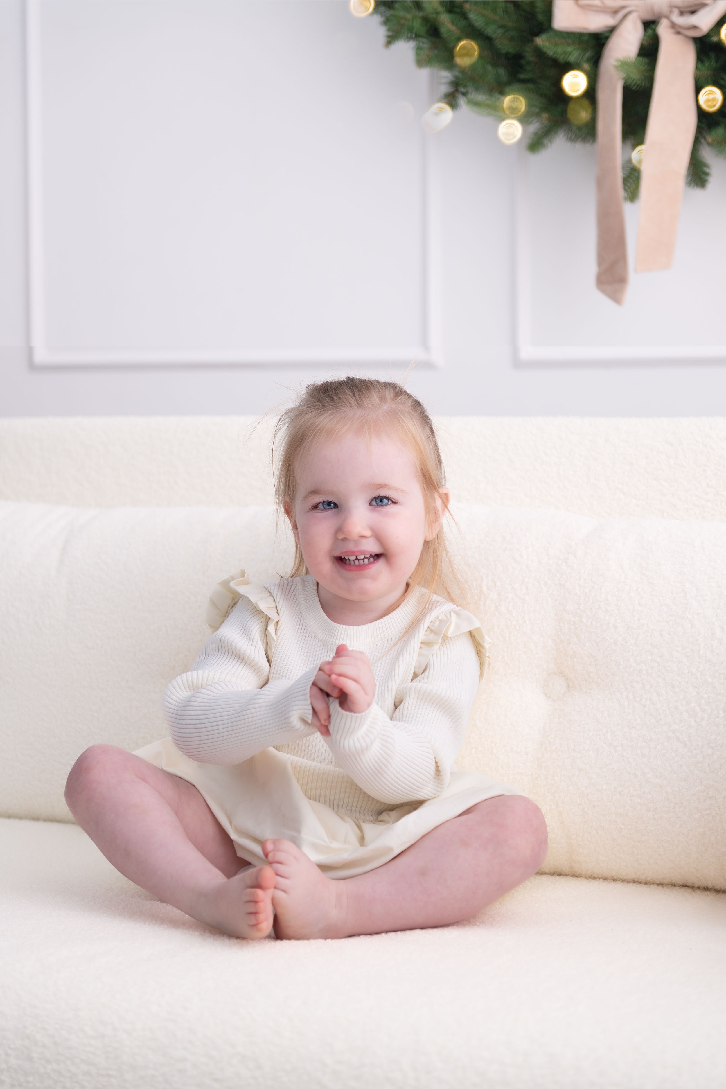 A small child wearing a cream colored sweater sits on a white couch with holiday decor visible in the background.