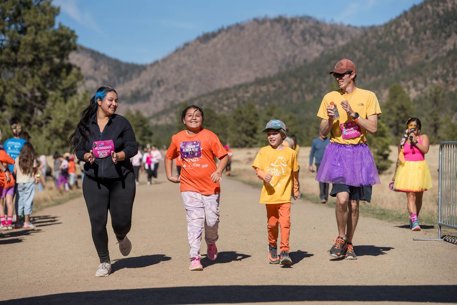 Colorfully dressed participants walk along a desert trail with mountains in the background during an outdoor event.