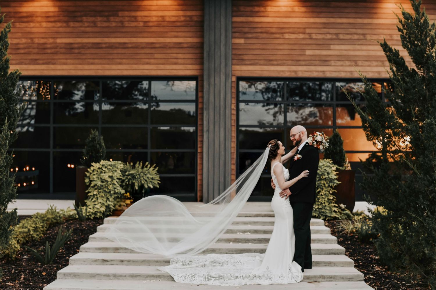 a husband and wife stand in front of each other and candidly hold each other as they smile and the bride's veil flows magically behind them