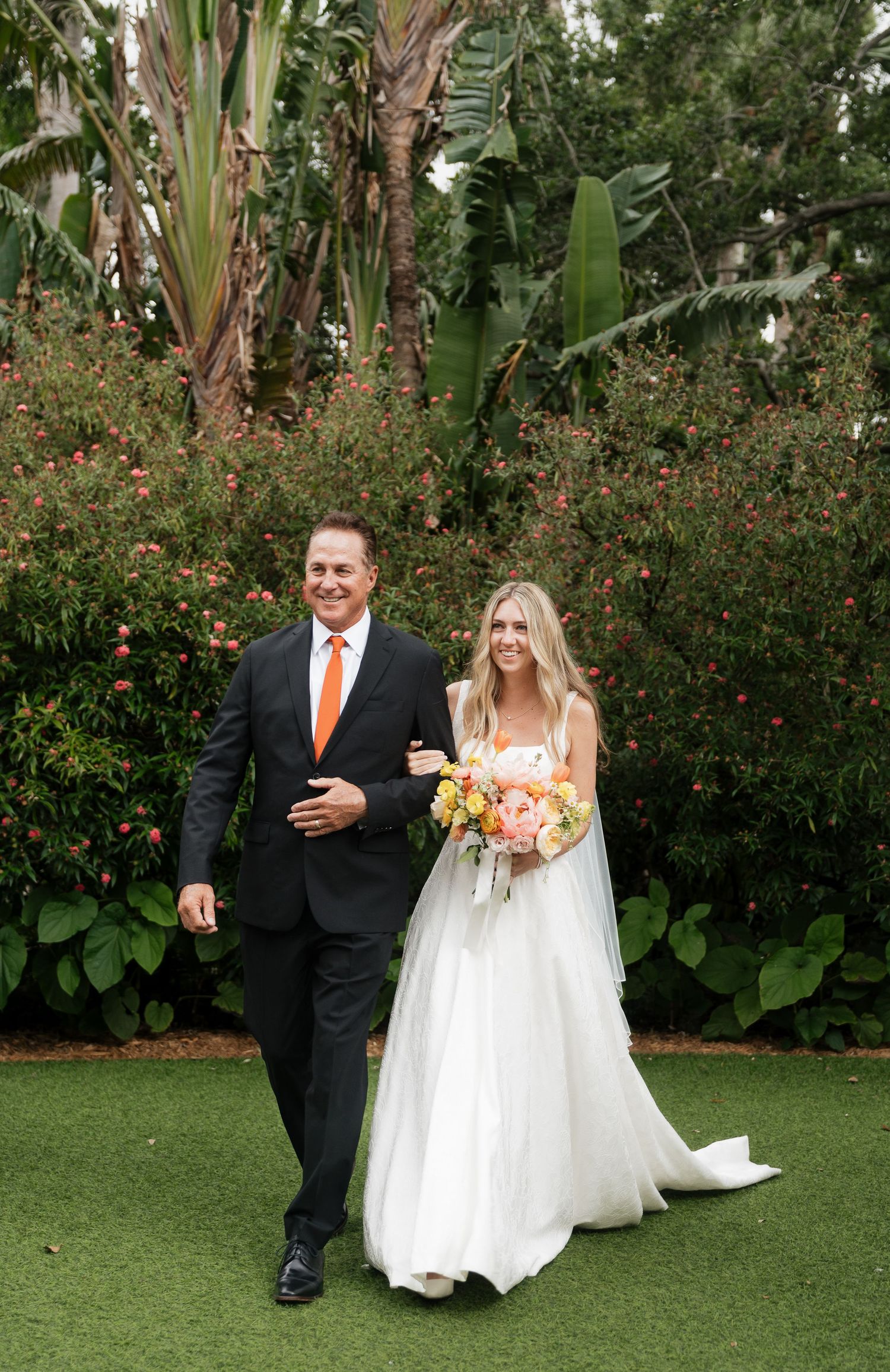 A wedding couple walks together on a lush green lawn with tropical foliage and red flowers in the background.