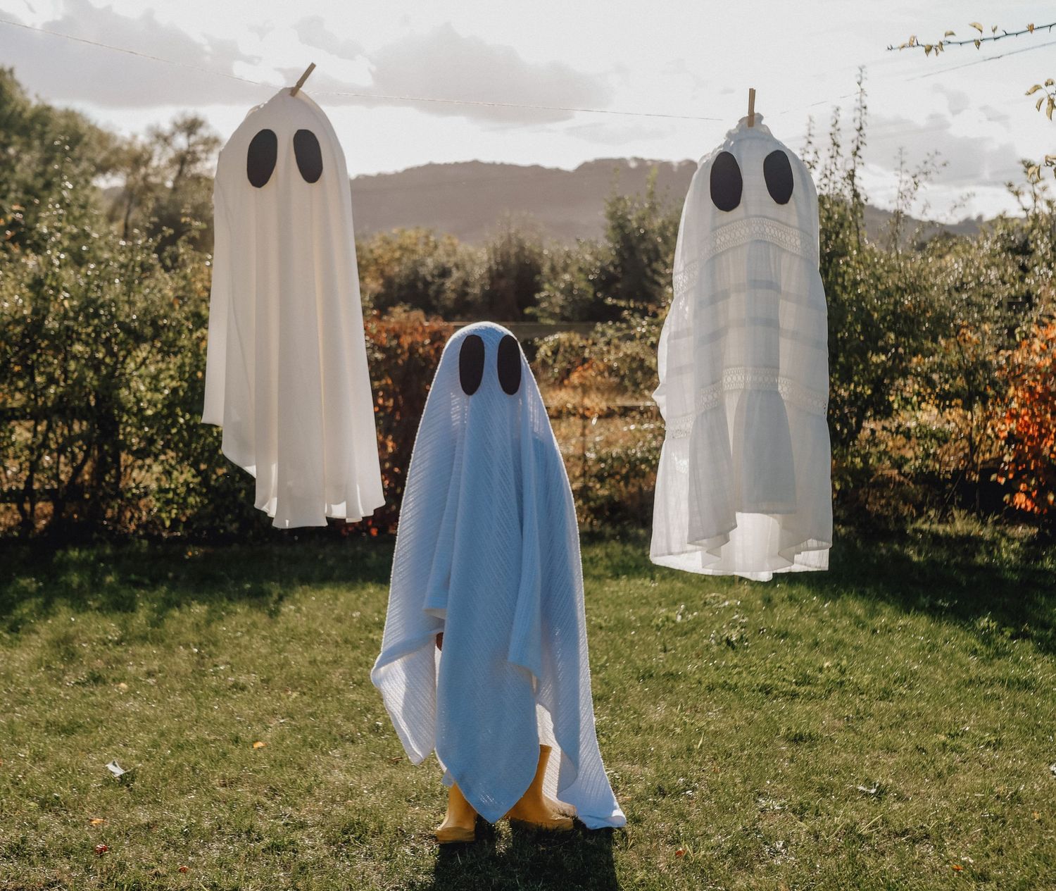 Five ghost decorations made from white sheets and blue fabric stand in a grassy field during autumn season.