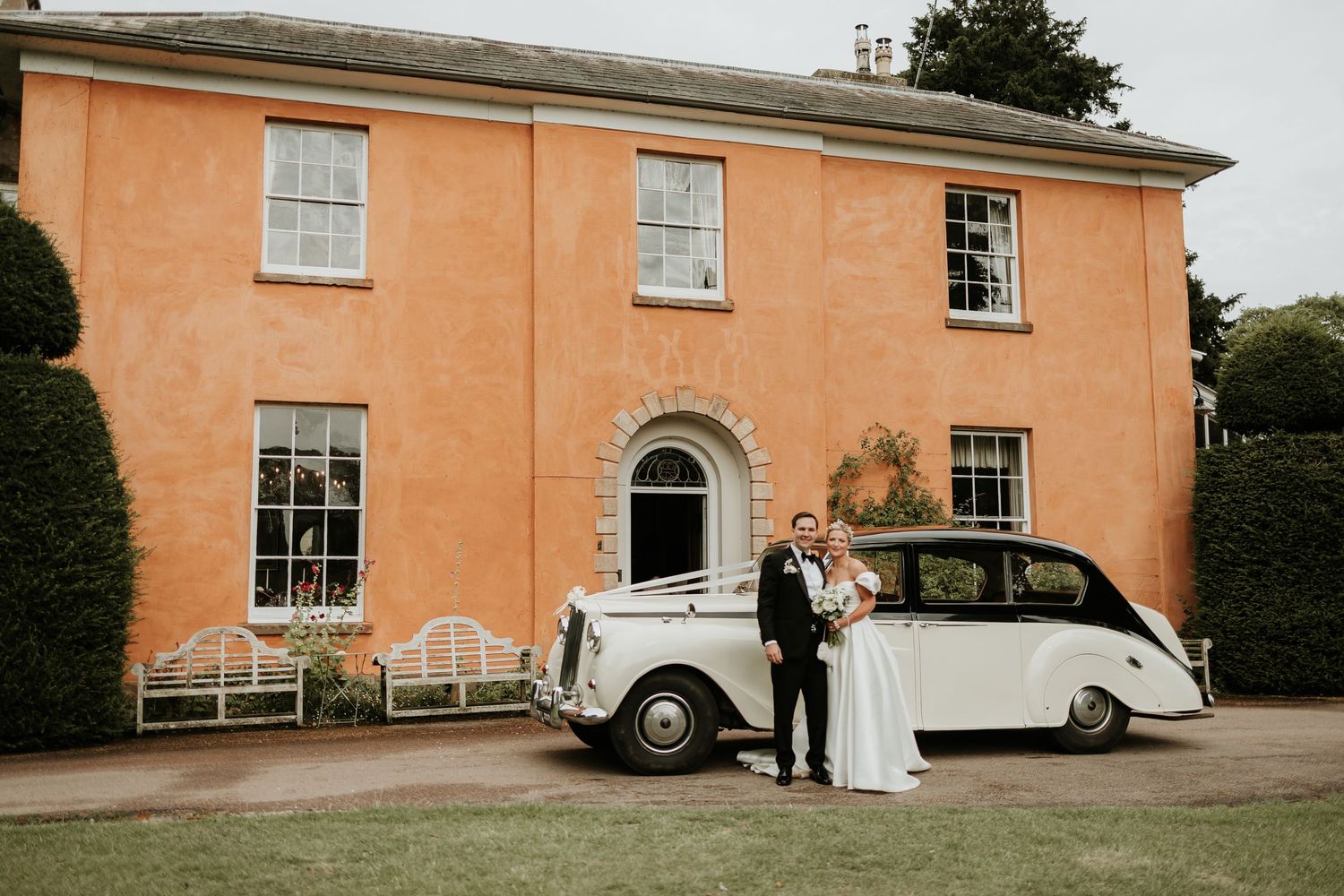A classic vintage car parked outside a peach-colored mansion during a wedding celebration.