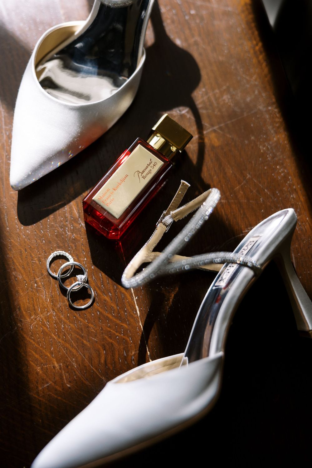 White bridal shoes, wedding rings and red perfume bottle arranged on a wooden surface in elegant composition.