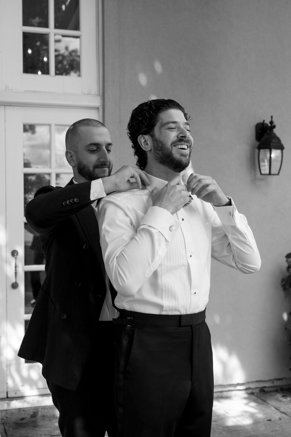 Black and white photo of groomsman helping groom adjust his bowtie while both laugh during wedding preparations.