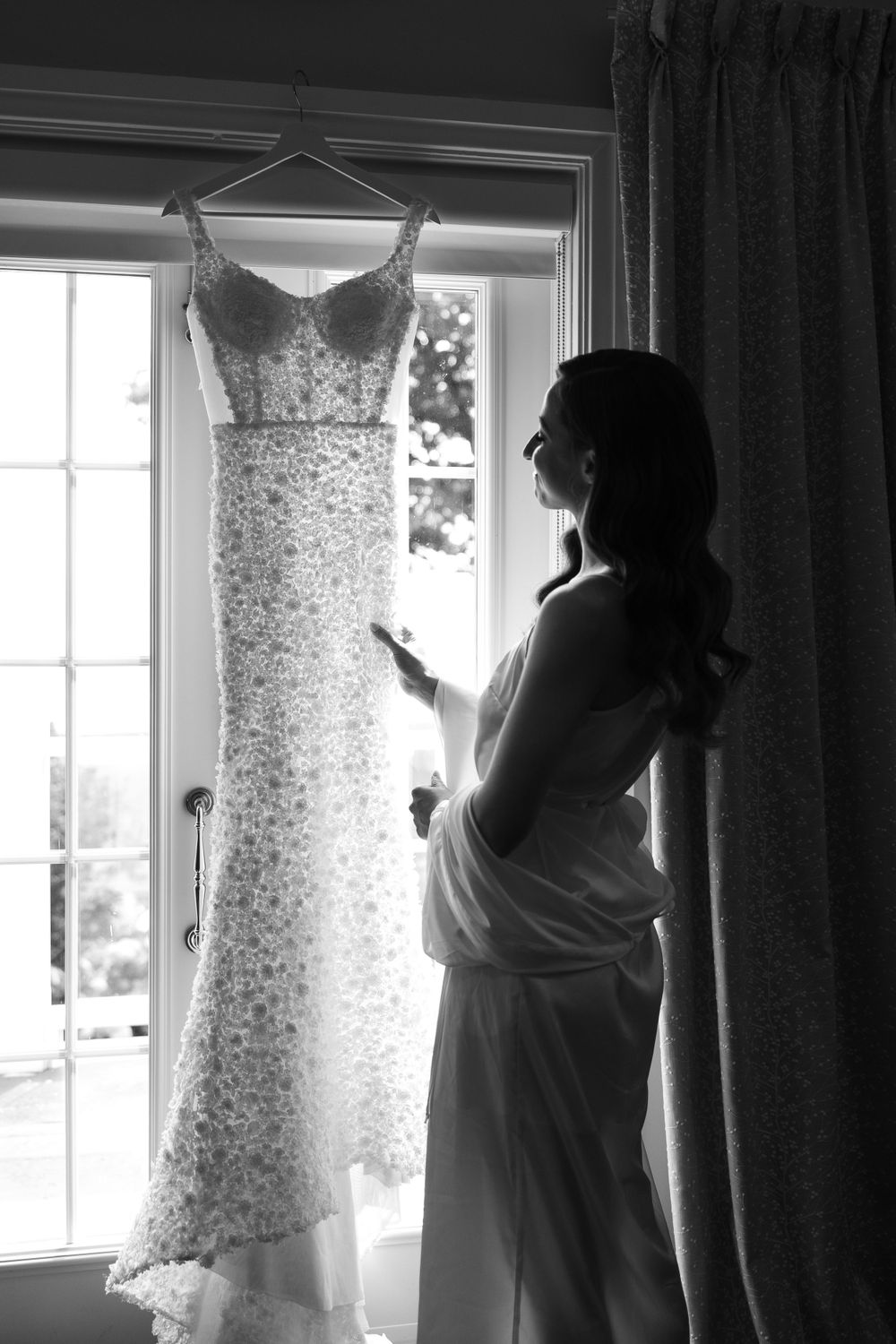 A silhouetted figure looks at a delicate lace wedding gown hanging in a window, creating a romantic black and white moment.