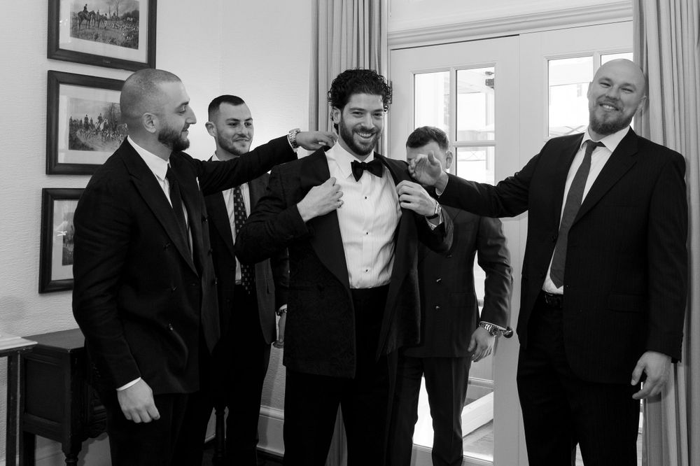 A black and white image of groomsmen adjusting a bowtie in a room with framed artwork on the wall.