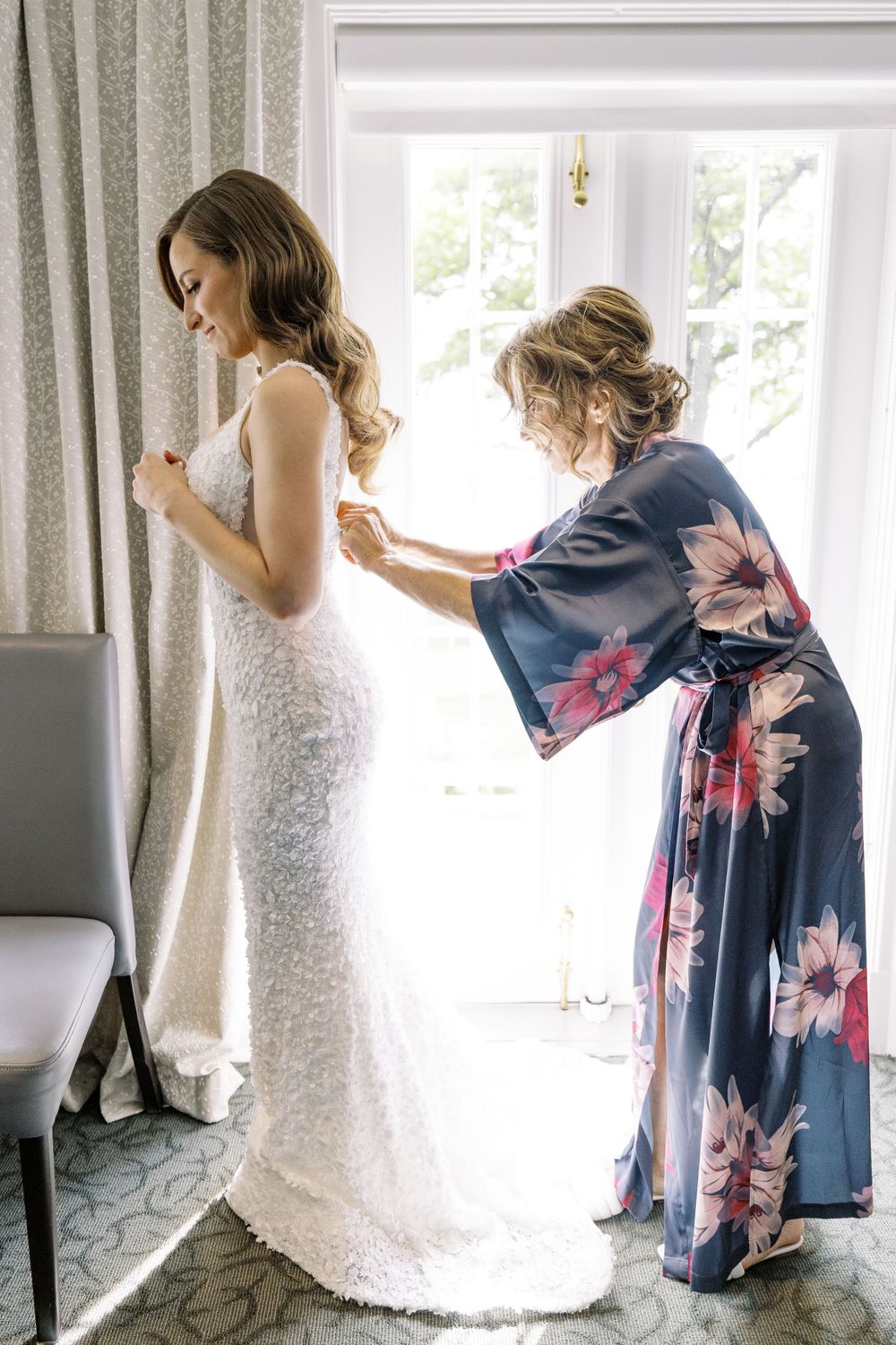 Helper in a floral robe assists with fastening an elegant white lace wedding gown in a sunlit room.