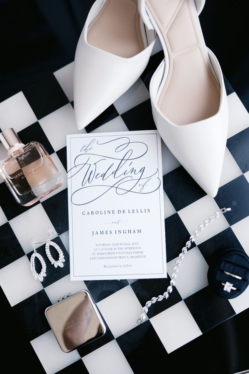Wedding invitation with elegant calligraphy displayed with white bridal shoes, perfume and jewelry on a black and white checkered surface.