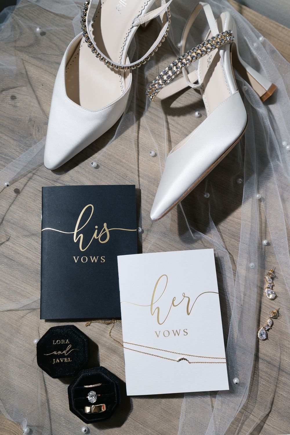 Wedding vow books and white bridal shoes arranged with jewelry on a light wooden surface.