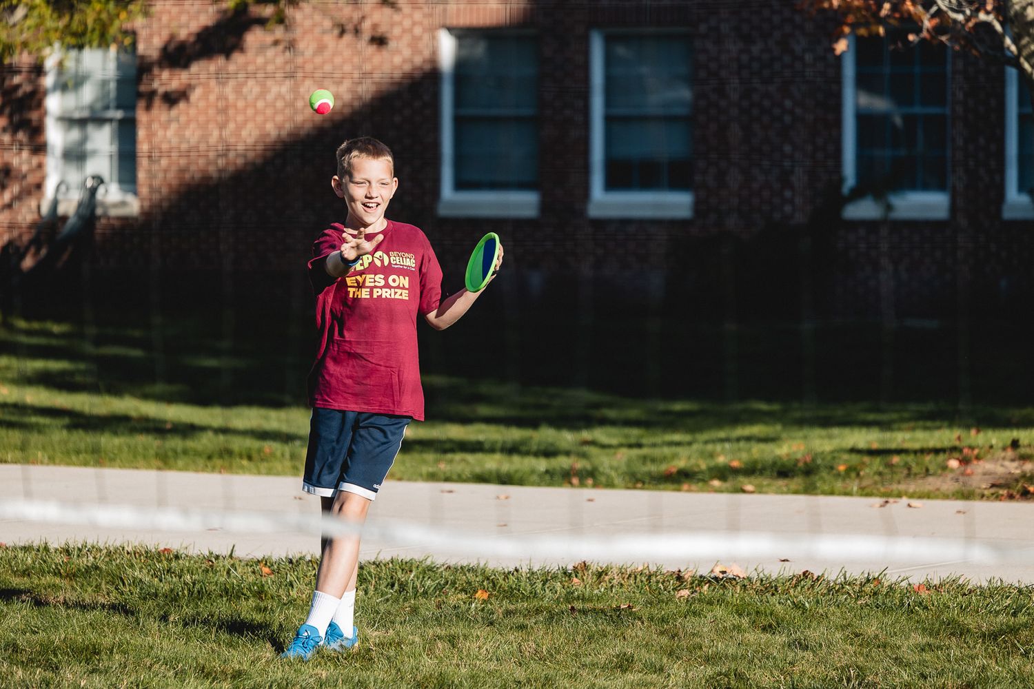A person in a red shirt plays with a soccer ball on a sunny grassy field outside a building.
