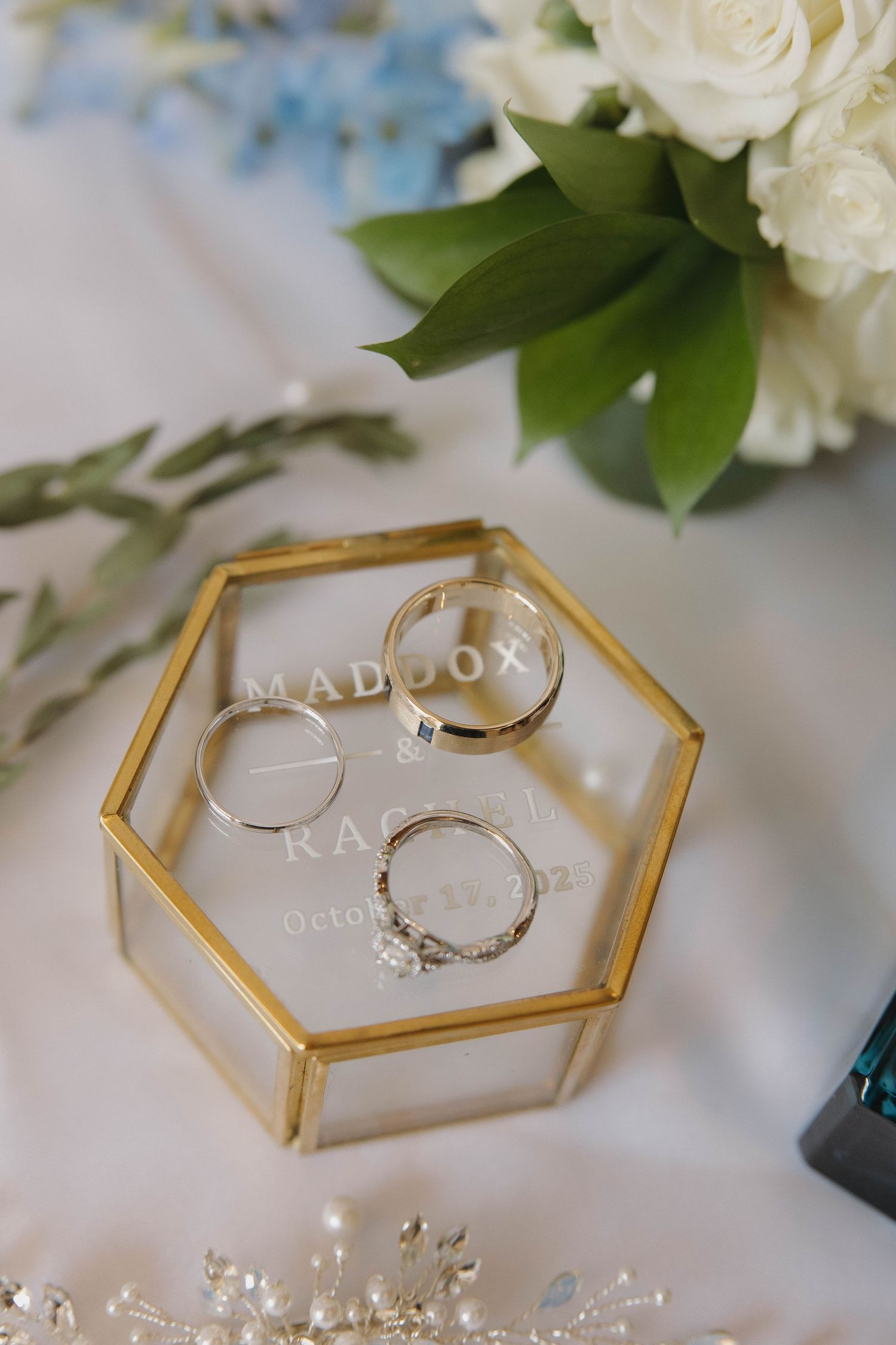 Wedding rings displayed in a gold hexagonal glass box with white roses and greenery in the background.
