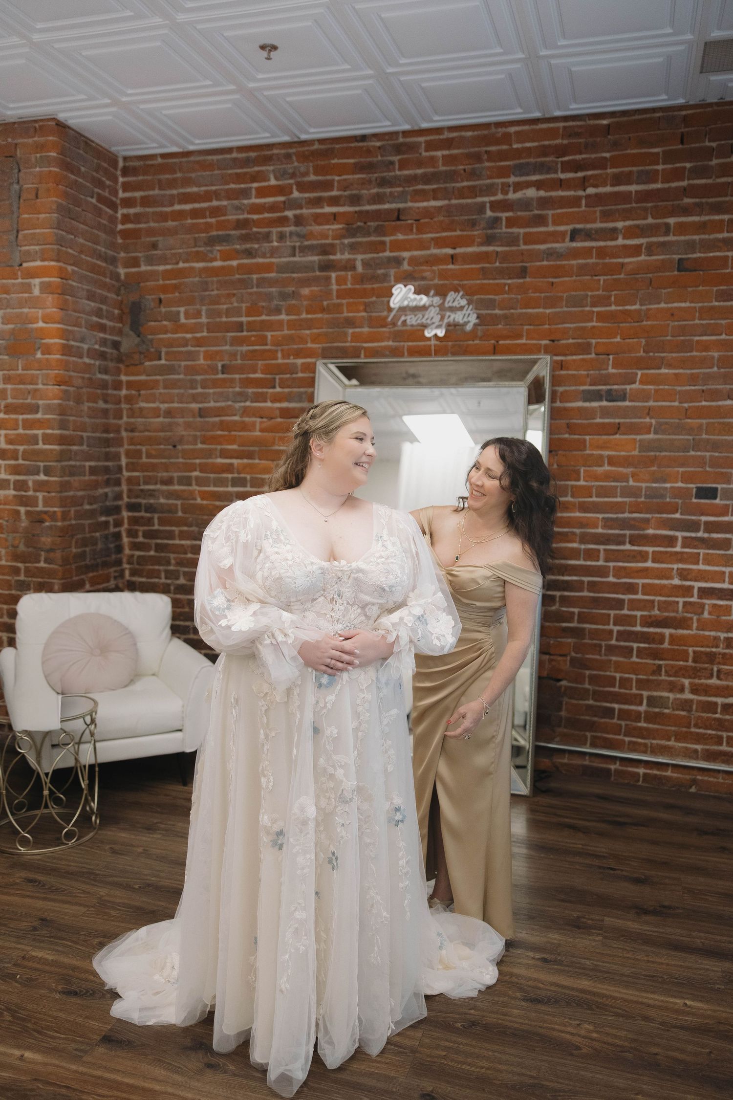 Two women in formal dresses share a joyful moment in a room with exposed brick walls and hardwood floors.