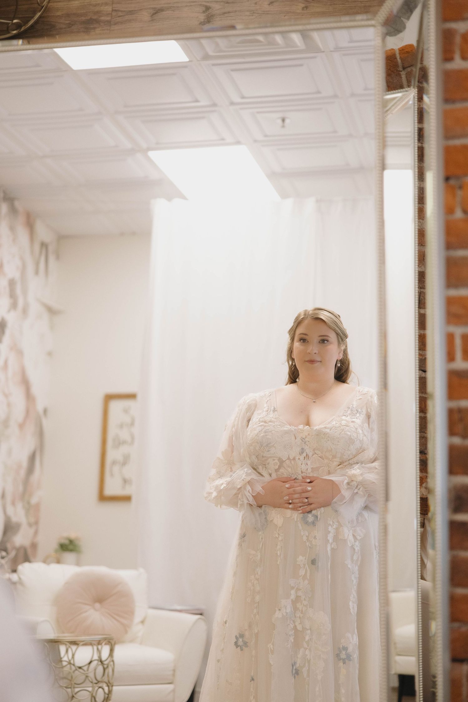 A bride in an elegant lace wedding dress poses in a bright white interior room with natural lighting.
