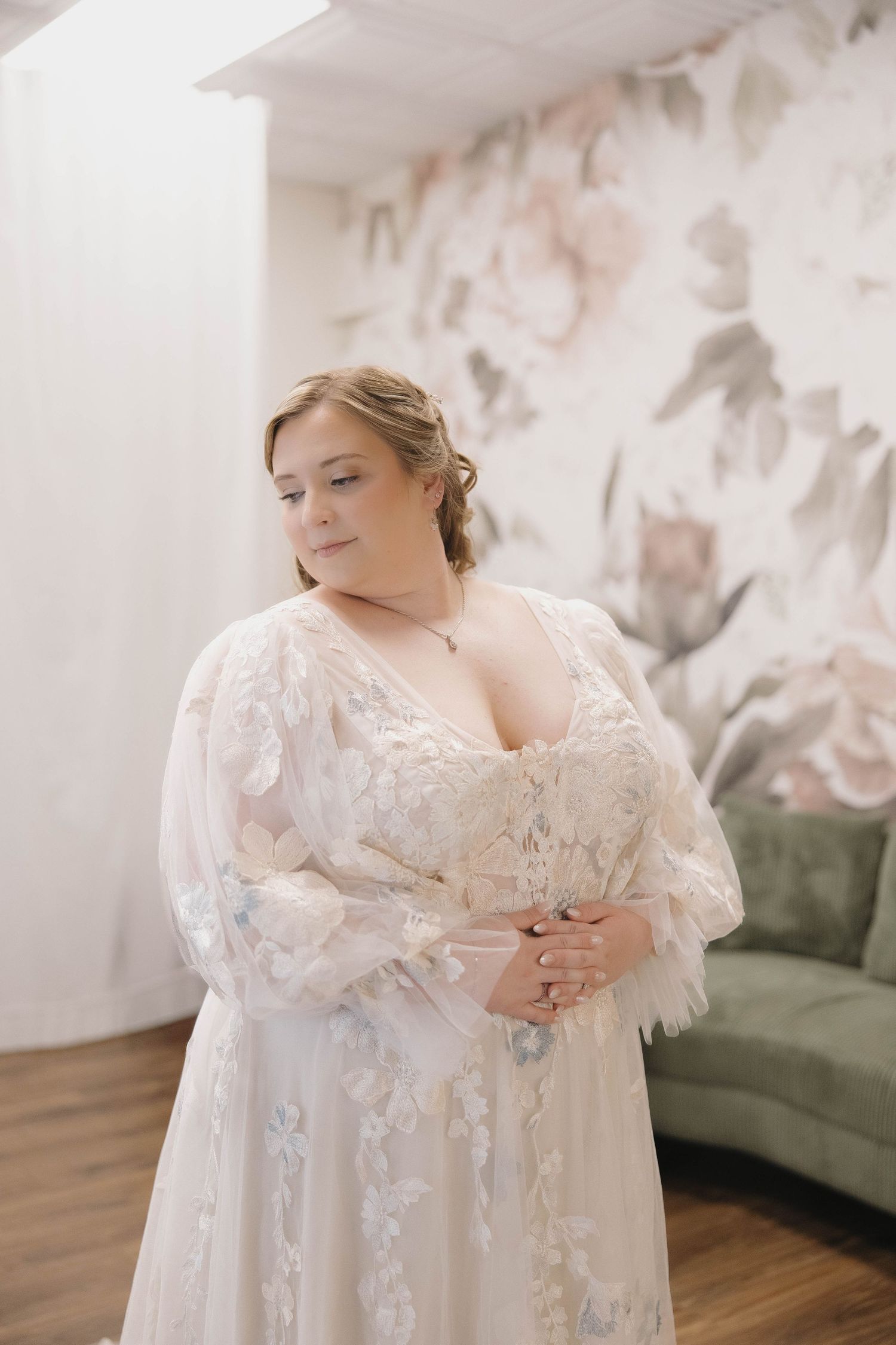 Elegant bride wearing a flowing white lace gown with feather details poses in a light-filled room with floral wallpaper.