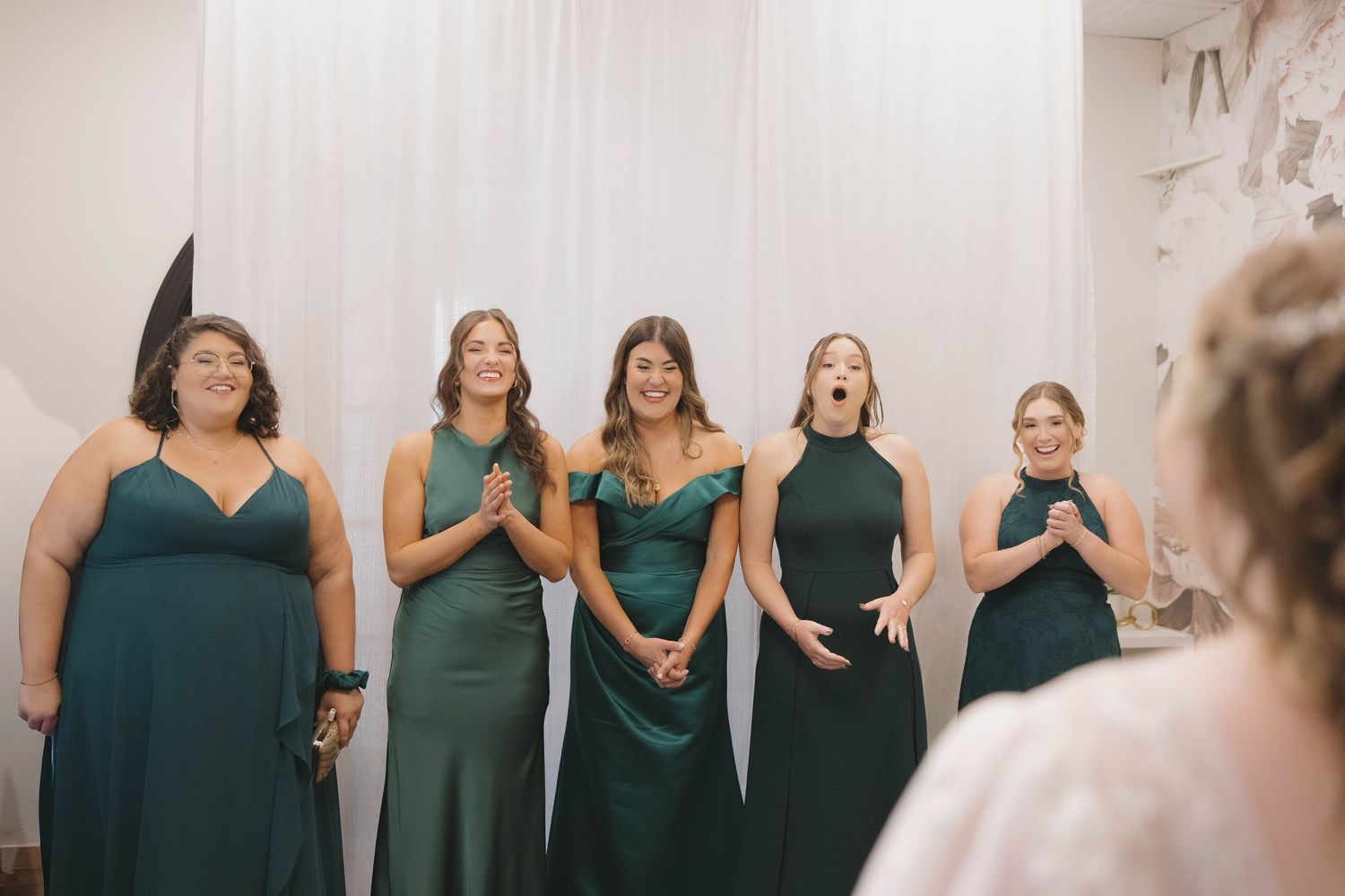 A row of bridesmaids wearing emerald green dresses stand together in a wedding venue.
