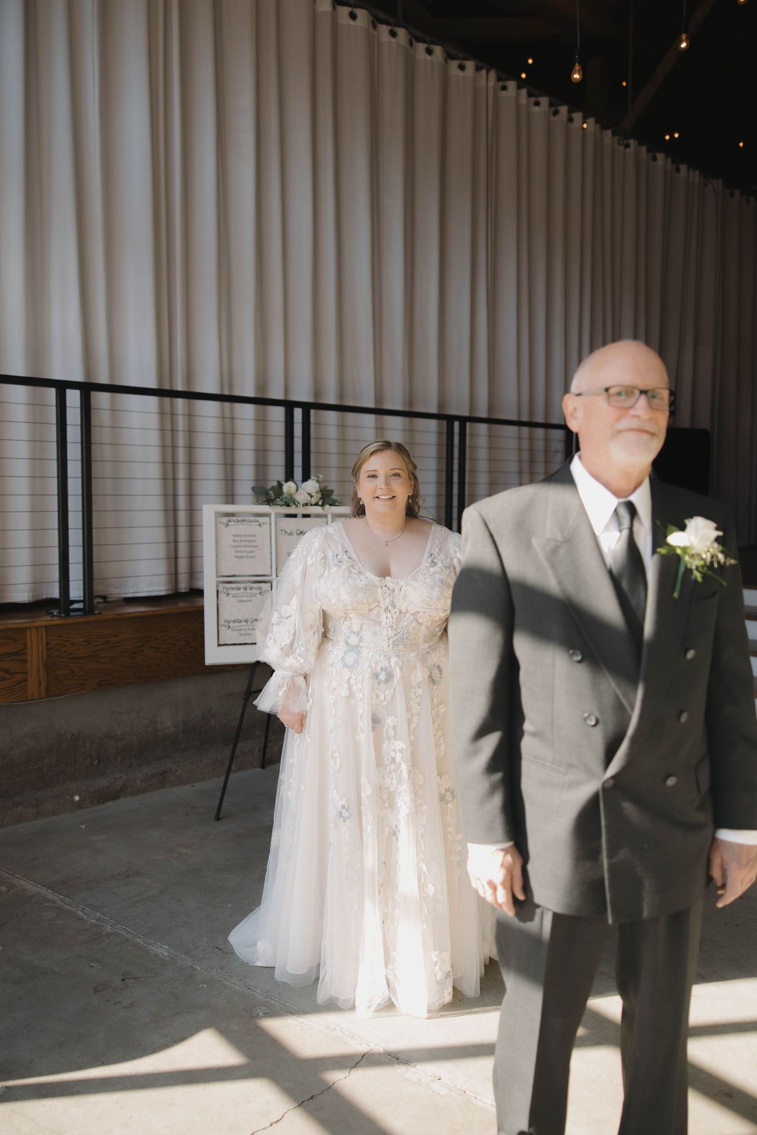 A couple in wedding attire stands in an industrial space with white curtains and dramatic lighting.