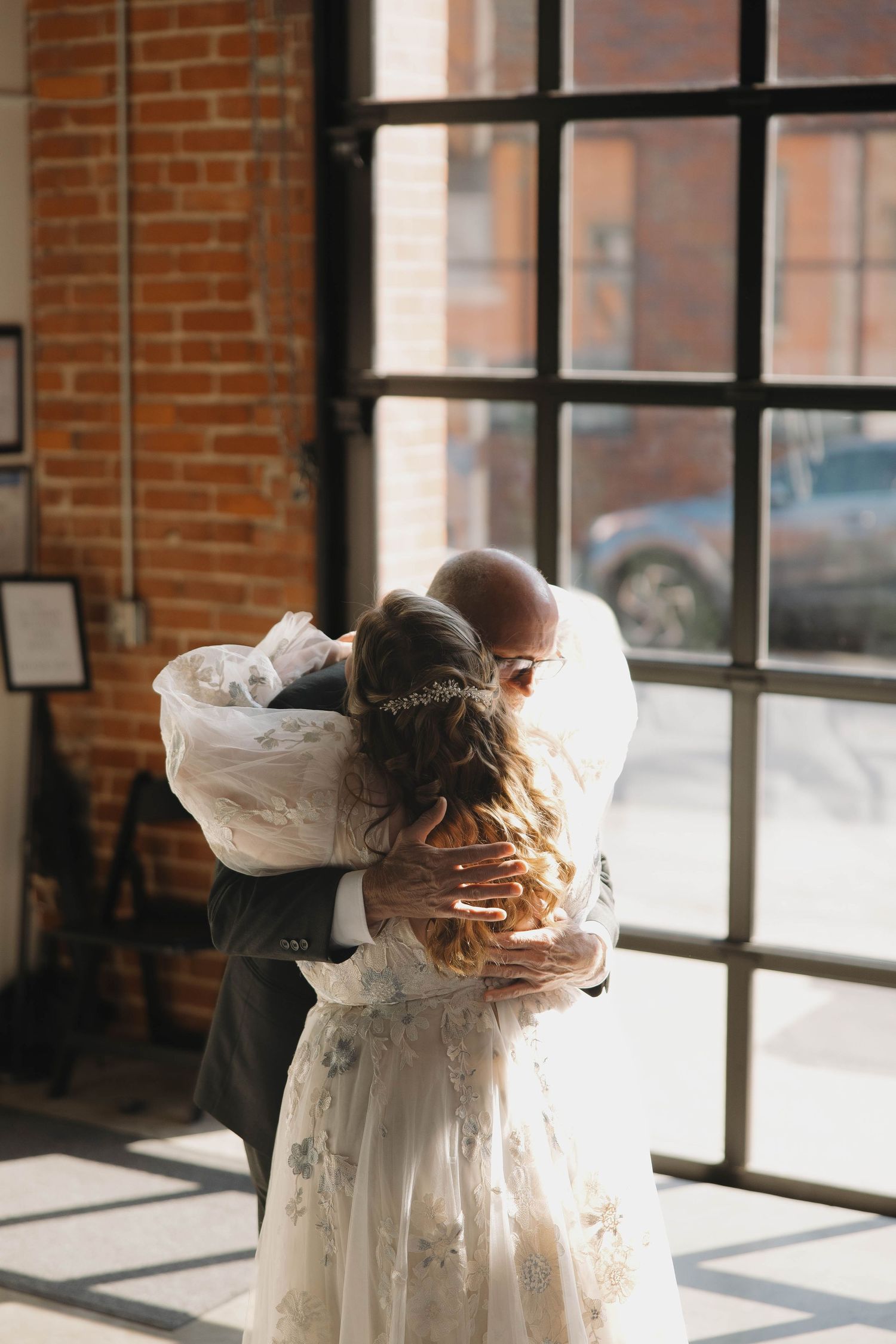 Two people embrace in an intimate moment by large windows in a brick-walled venue.
