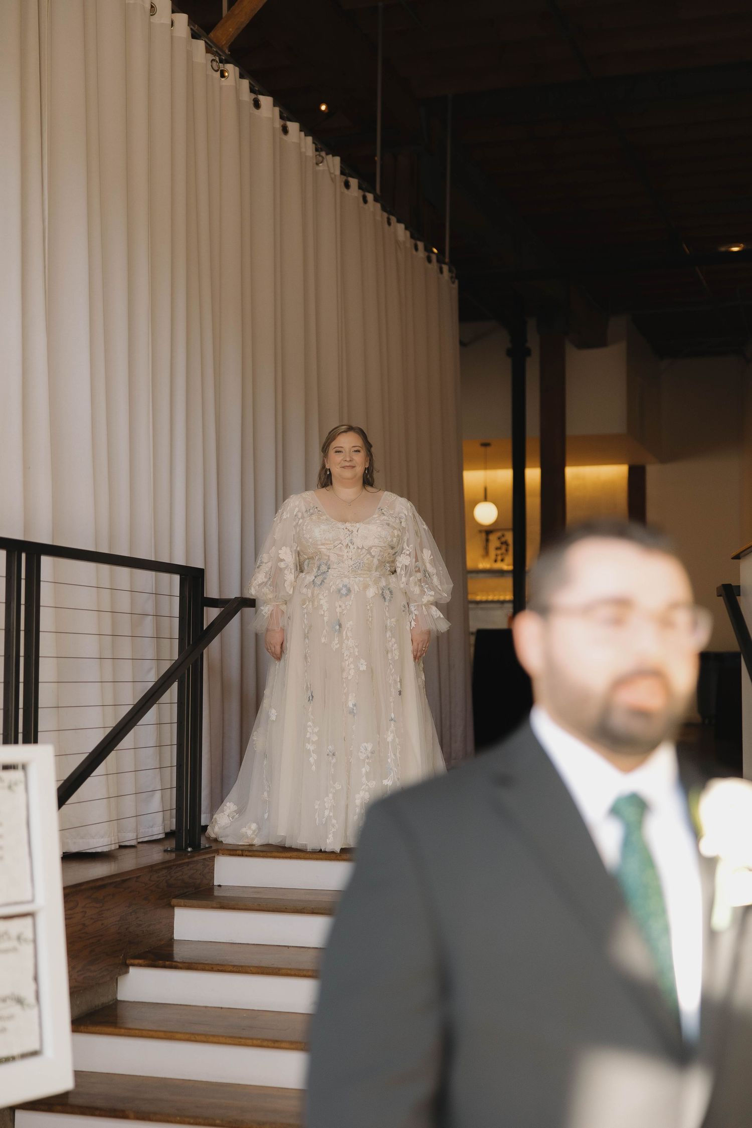 A bride in a sparkly white wedding gown stands at the top of white stairs while a man in a suit stands in the foreground.