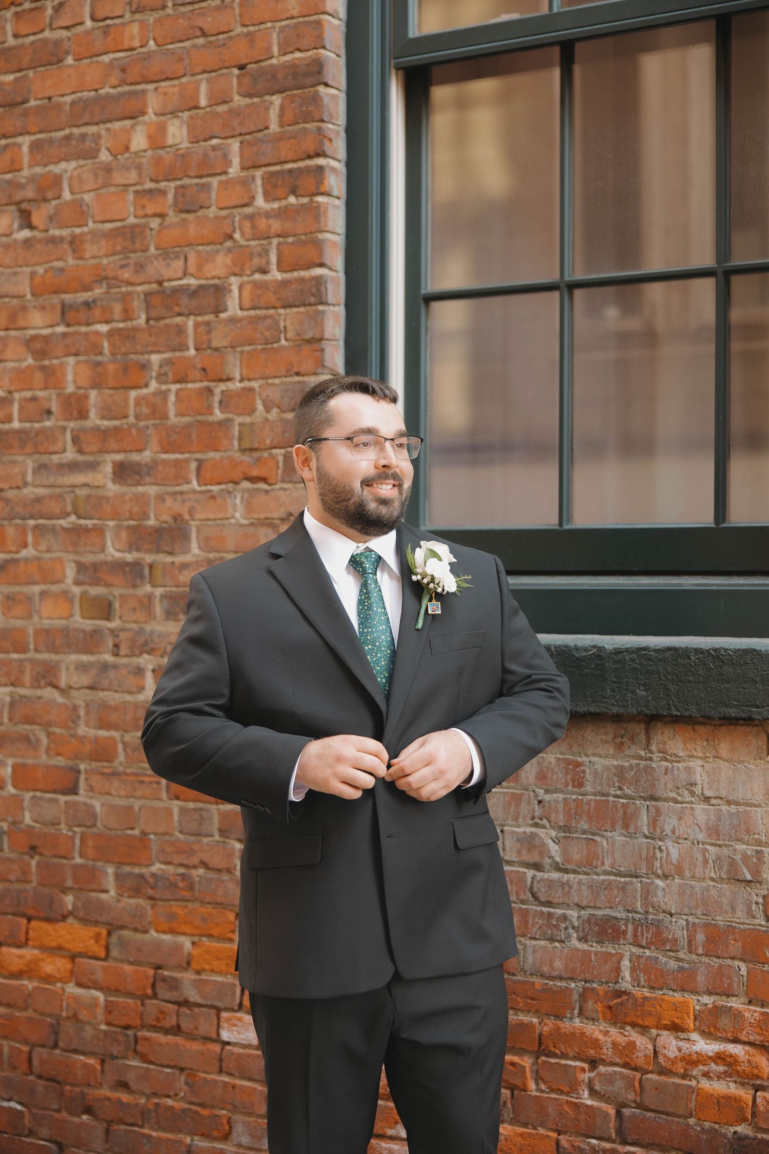 A groom in a black suit and teal tie poses against a rustic brick wall exterior.
