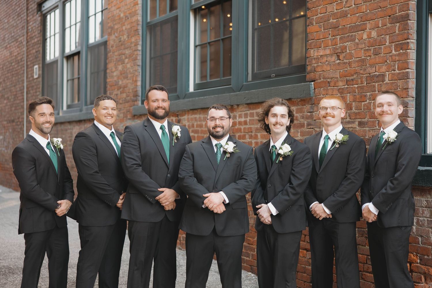 A wedding party in black suits and dresses poses in front of a rustic brick building.