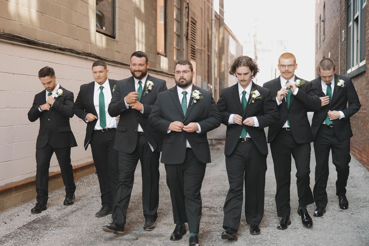 Wedding party in black suits poses playfully in urban alleyway with brick buildings in background.