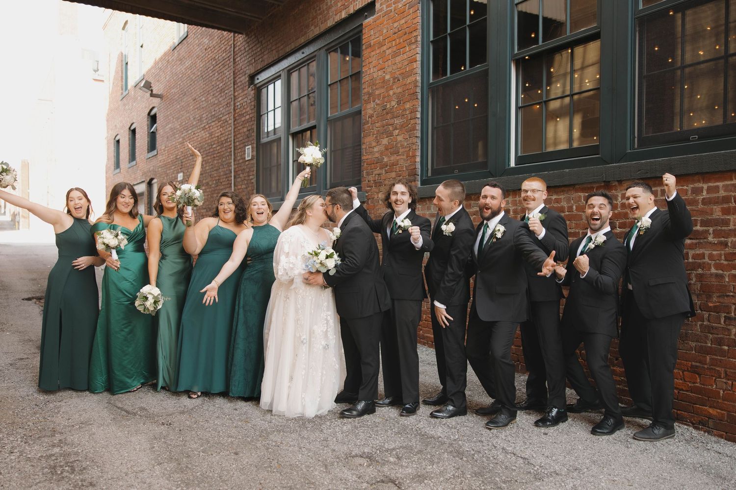 Wedding party in green dresses and black suits sits together against rustic brick wall backdrop.