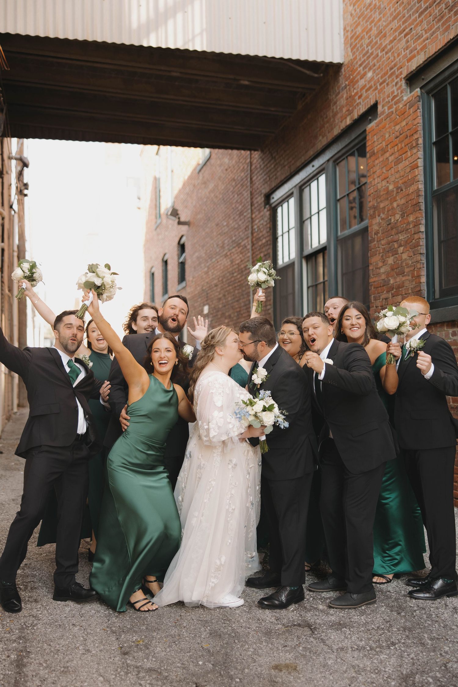 Wedding party poses playfully in an urban alley with brick walls, wearing black suits and emerald green dresses.