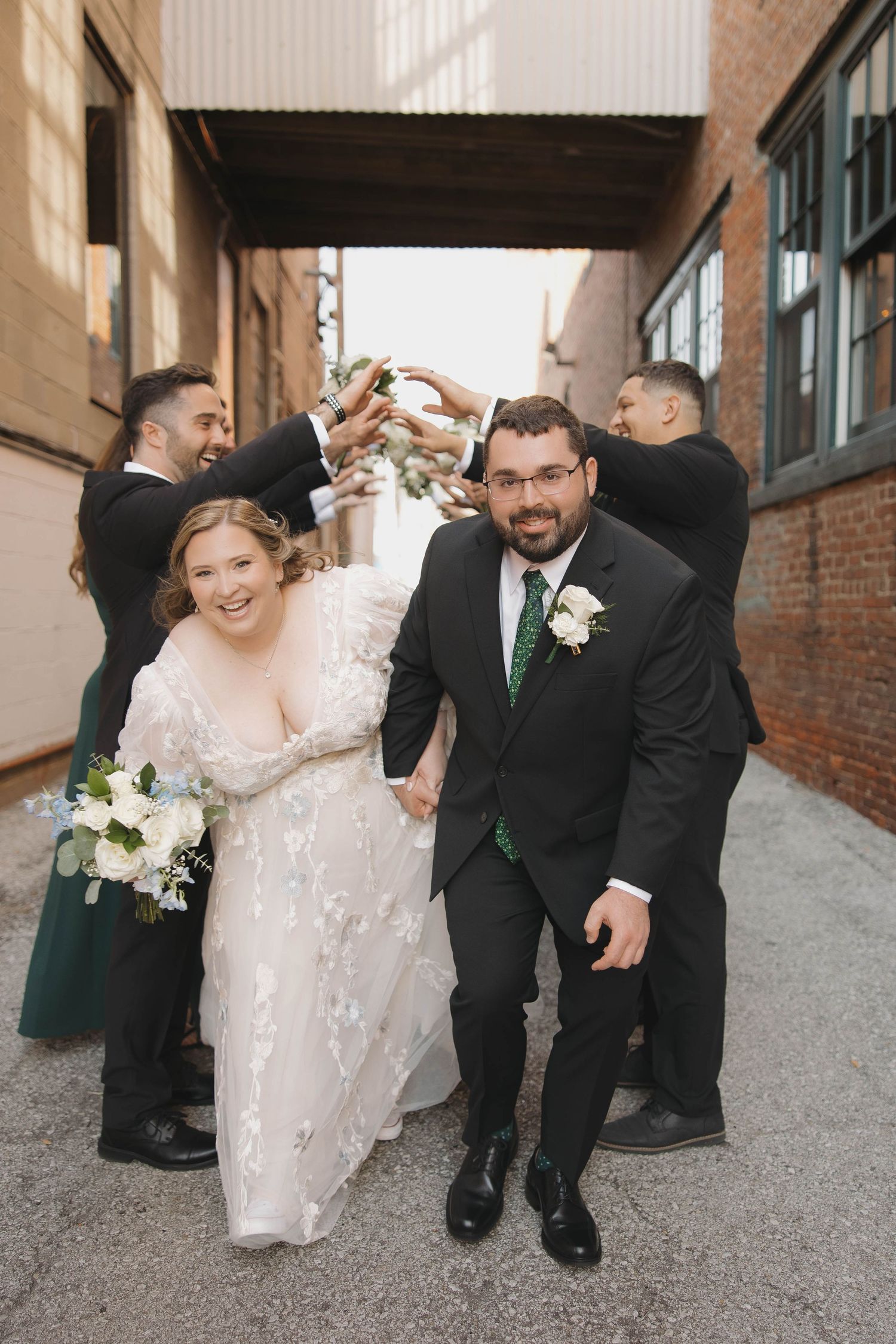 A wedding party walks and laughs together along a brick-lined alley in formal attire.