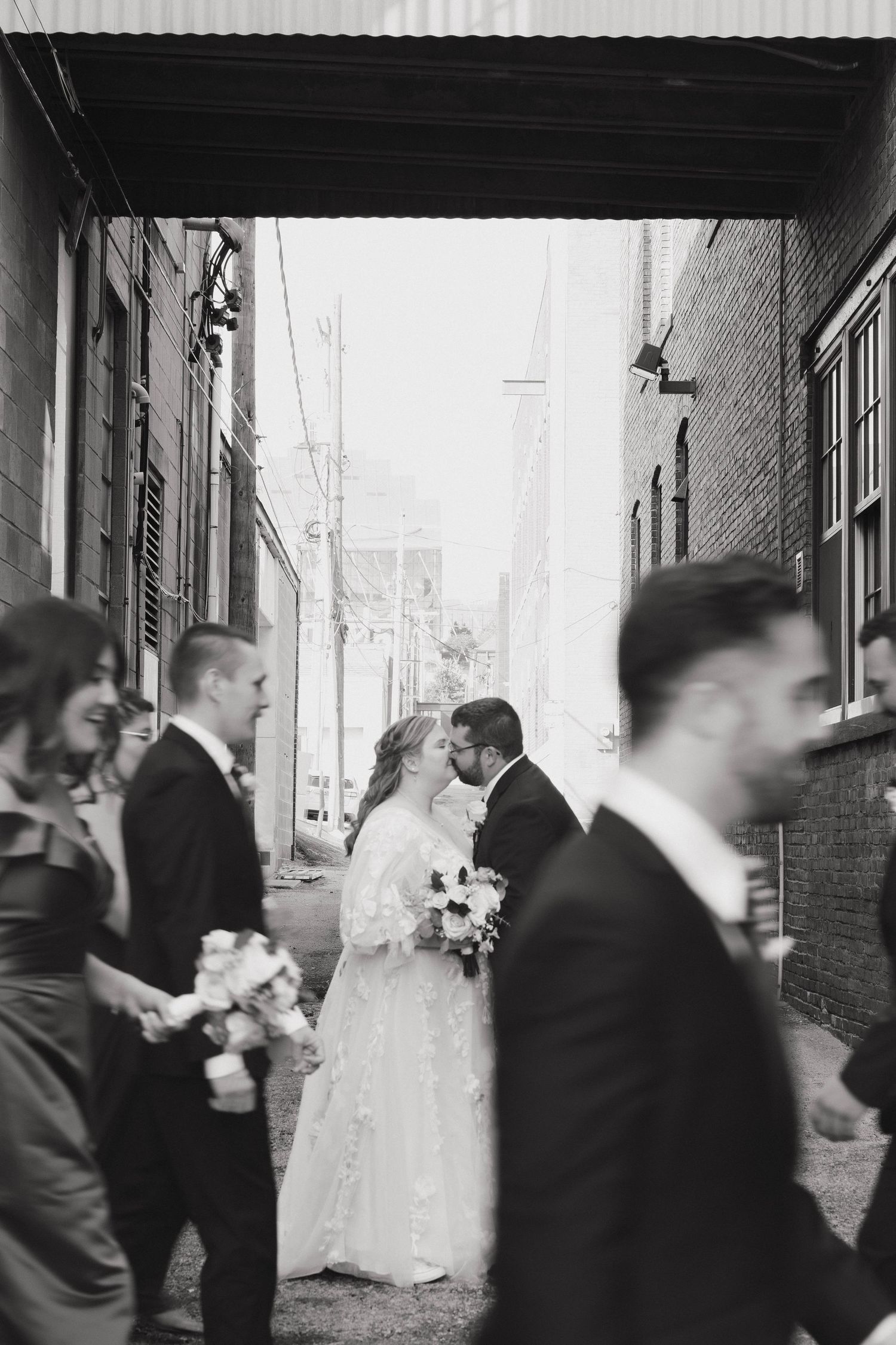 A black and white wedding party photo showing movement and candid moments along a brick building.