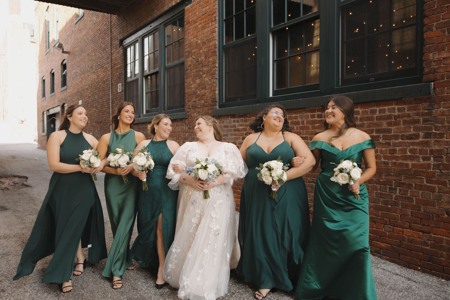 Wedding party members in emerald green dresses pose with the bride against a brick wall backdrop.