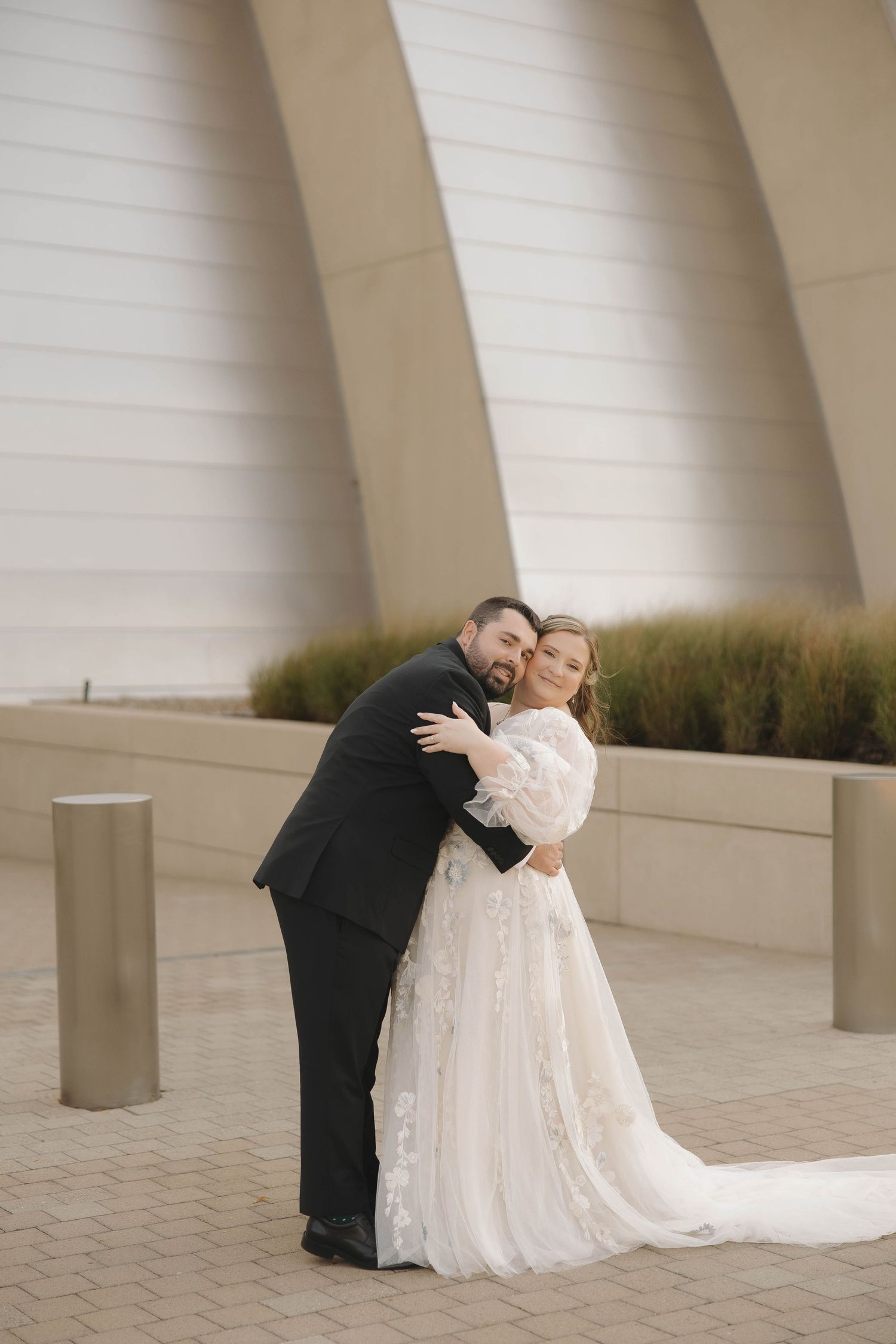 A couple in wedding attire embrace in front of a modern architectural backdrop with geometric white walls.