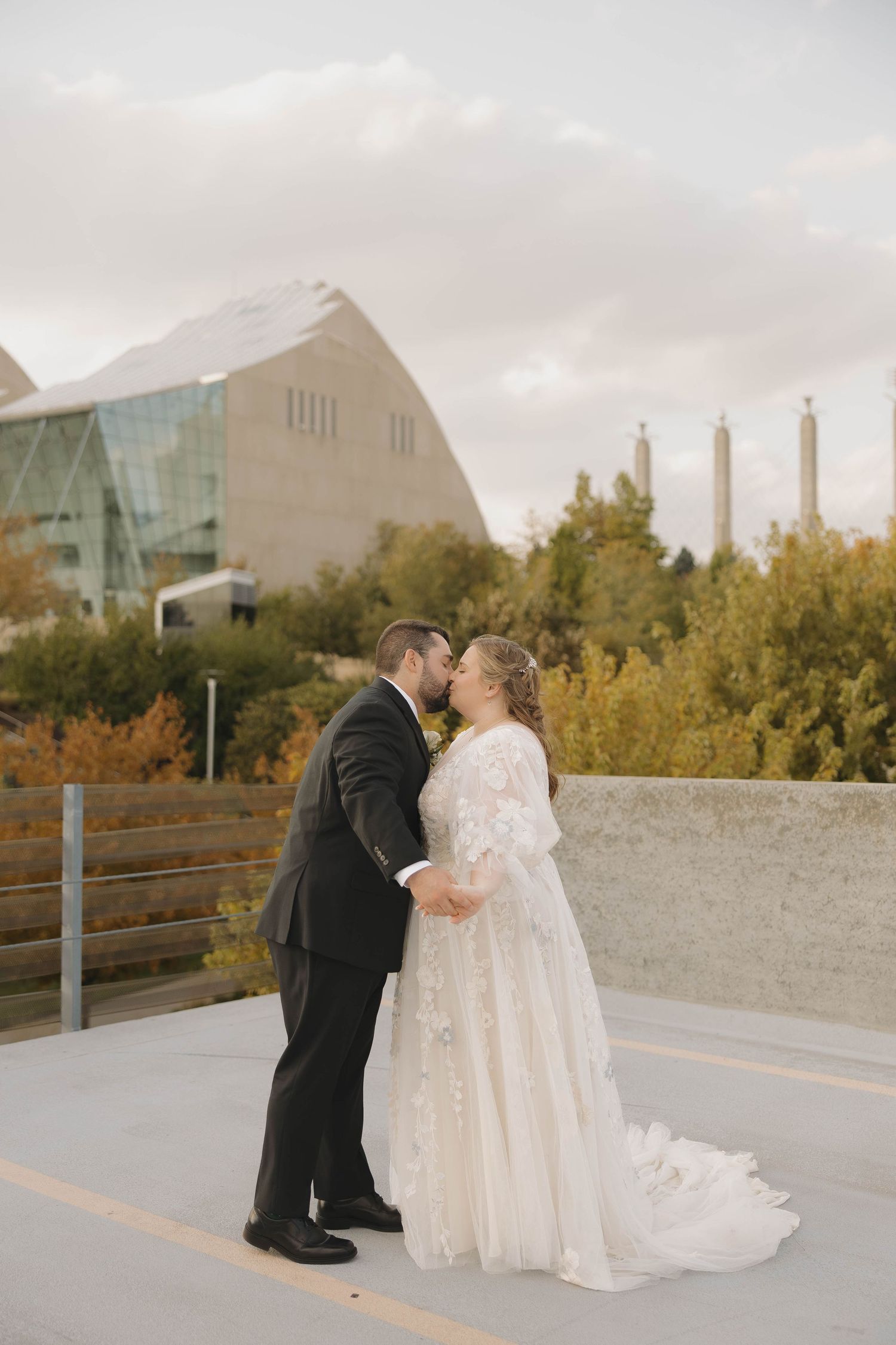 A couple in wedding attire share an intimate moment on a concrete walkway with modern buildings in the background.