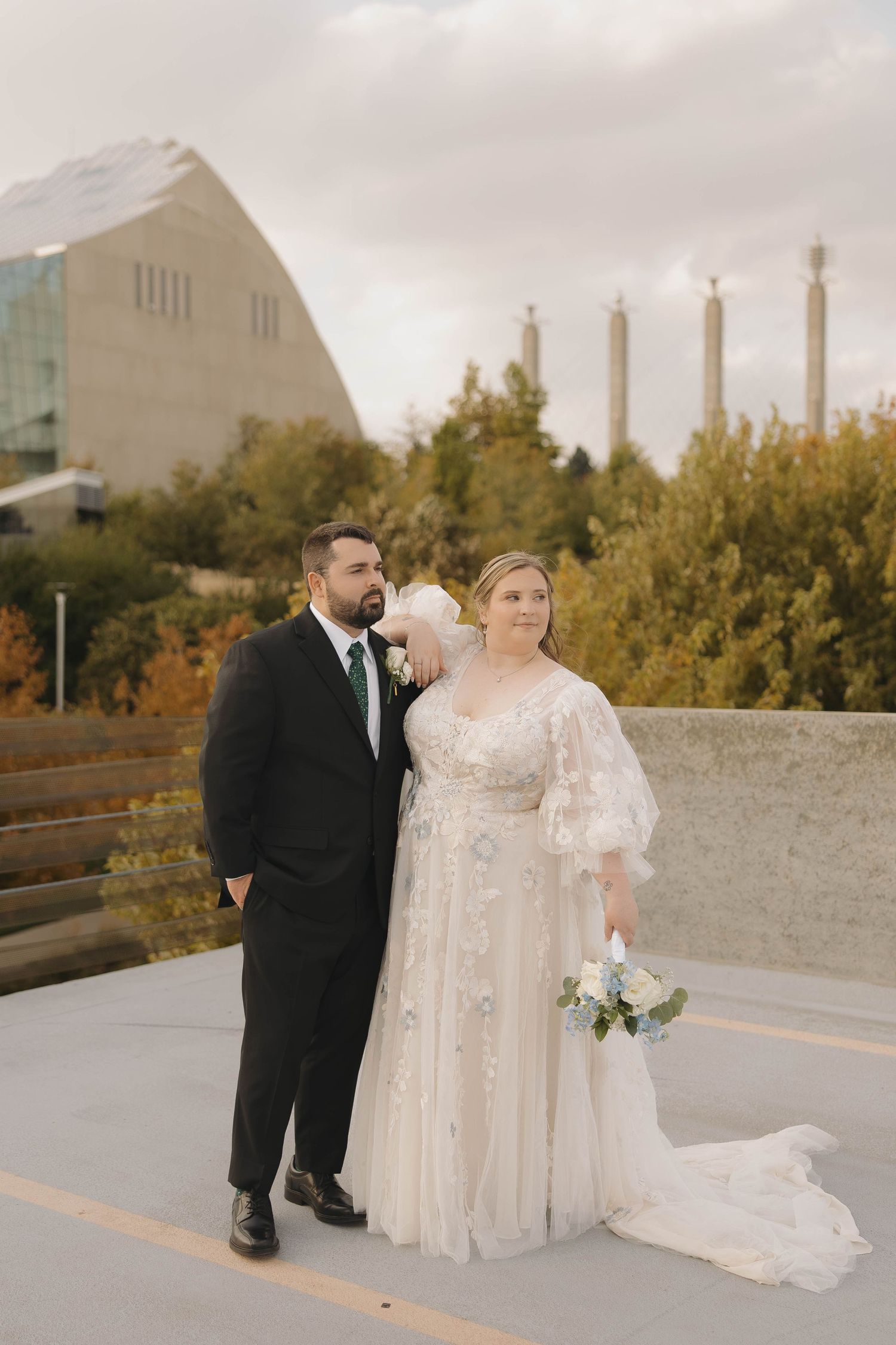 A couple poses for romantic wedding photos on a paved walkway surrounded by autumn trees and modern architecture.