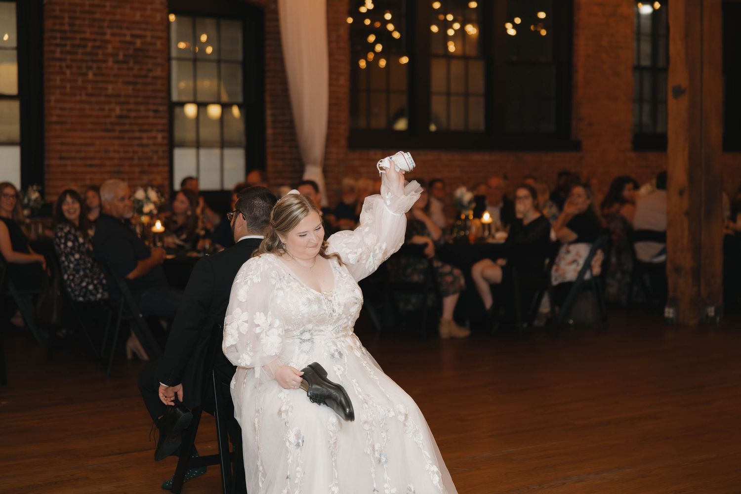 Wedding couple dances in a warmly lit brick venue with twinkling lights in the background.