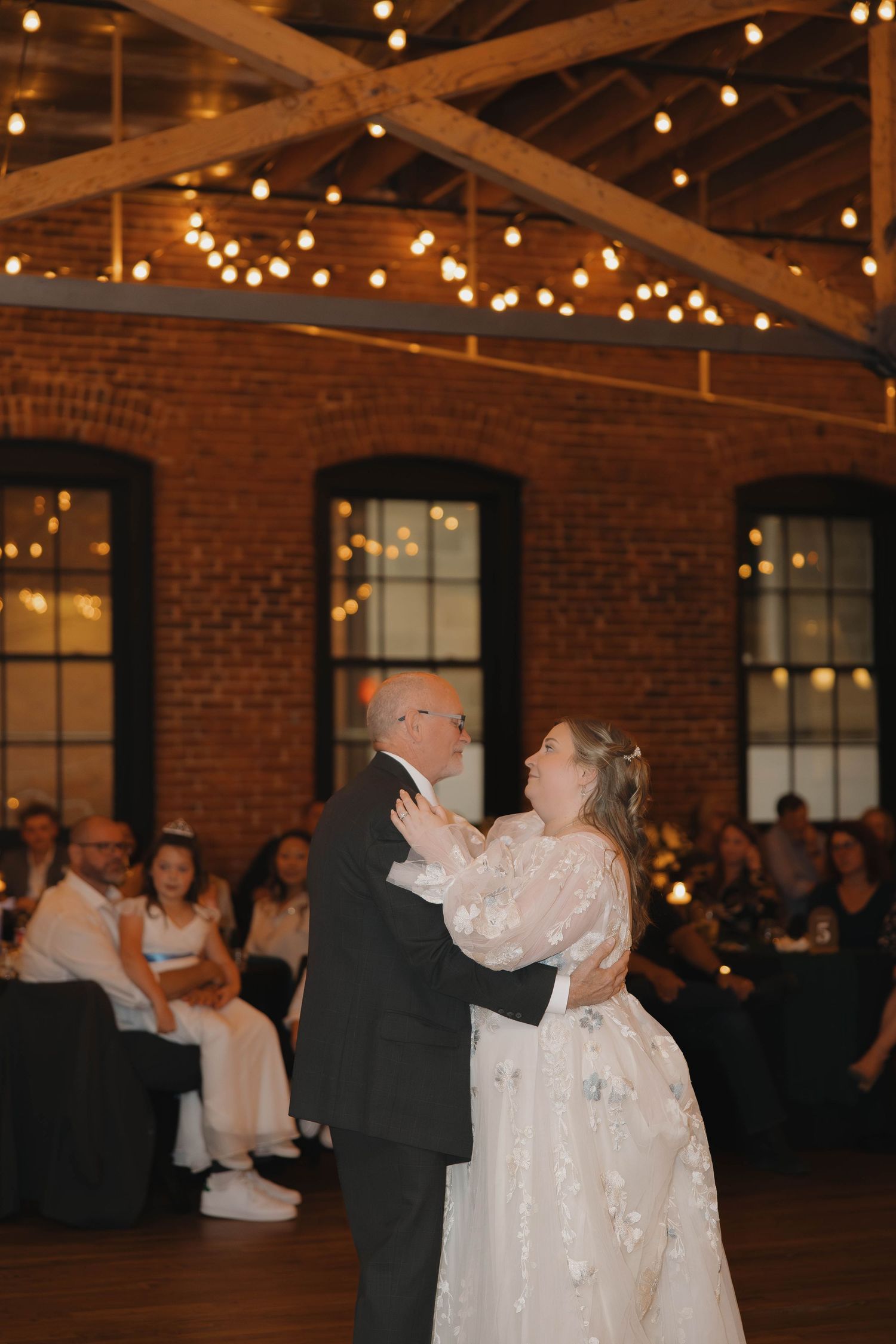 A couple dances together in a rustic brick venue decorated with romantic string lights overhead.