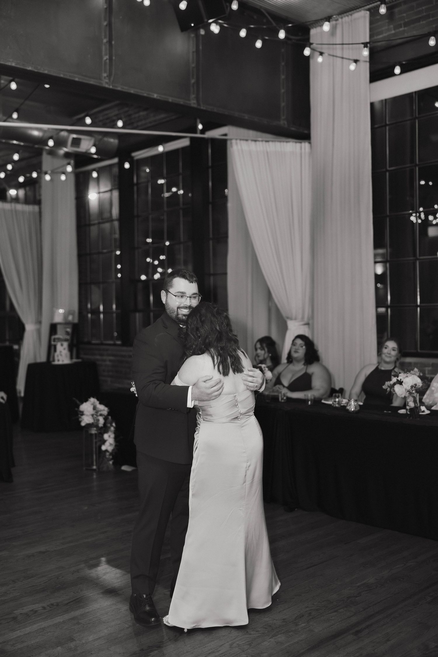 A couple dances in formal attire under string lights at an indoor reception venue.