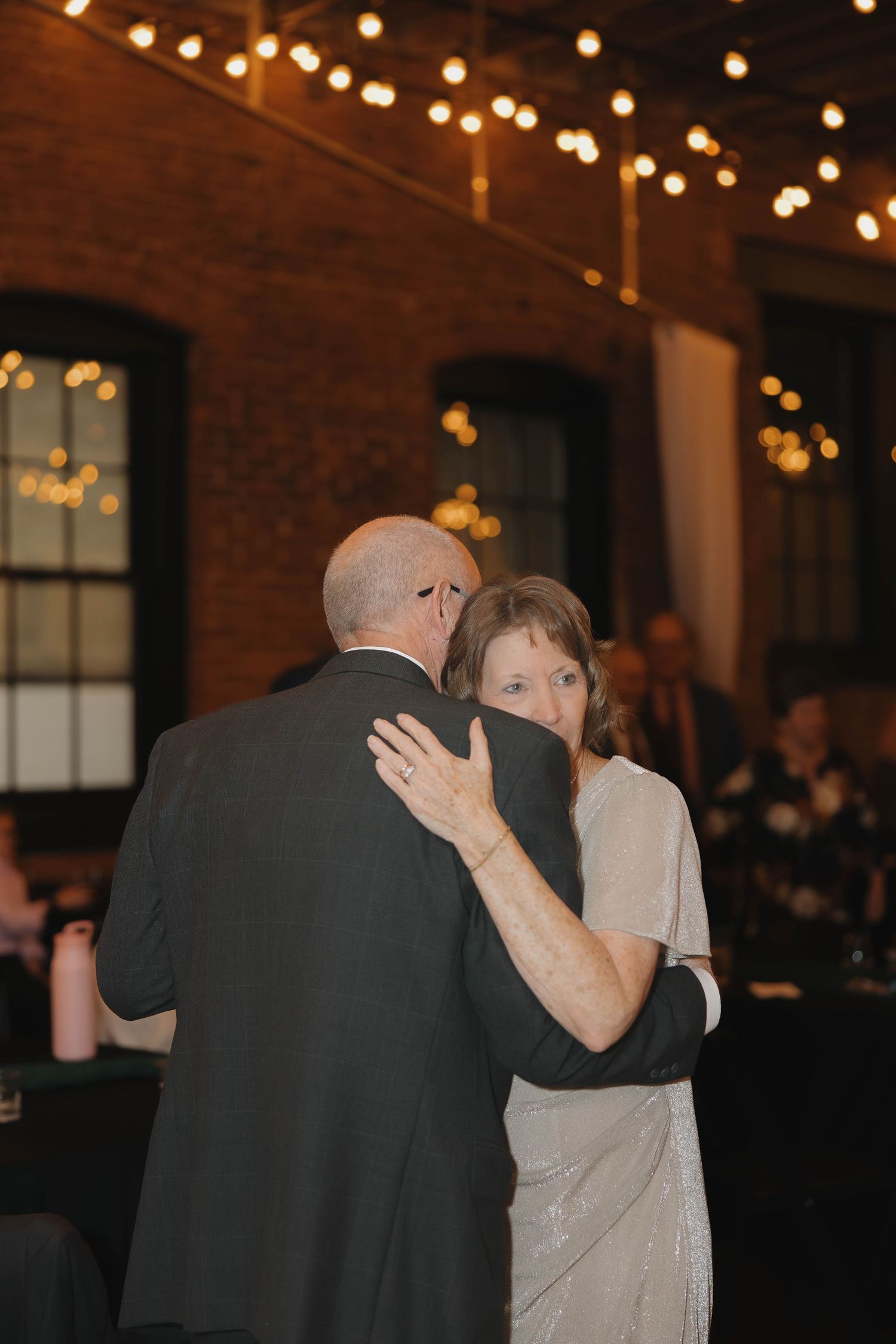 Wedding guests dance together under warm string lights in a rustic brick venue.