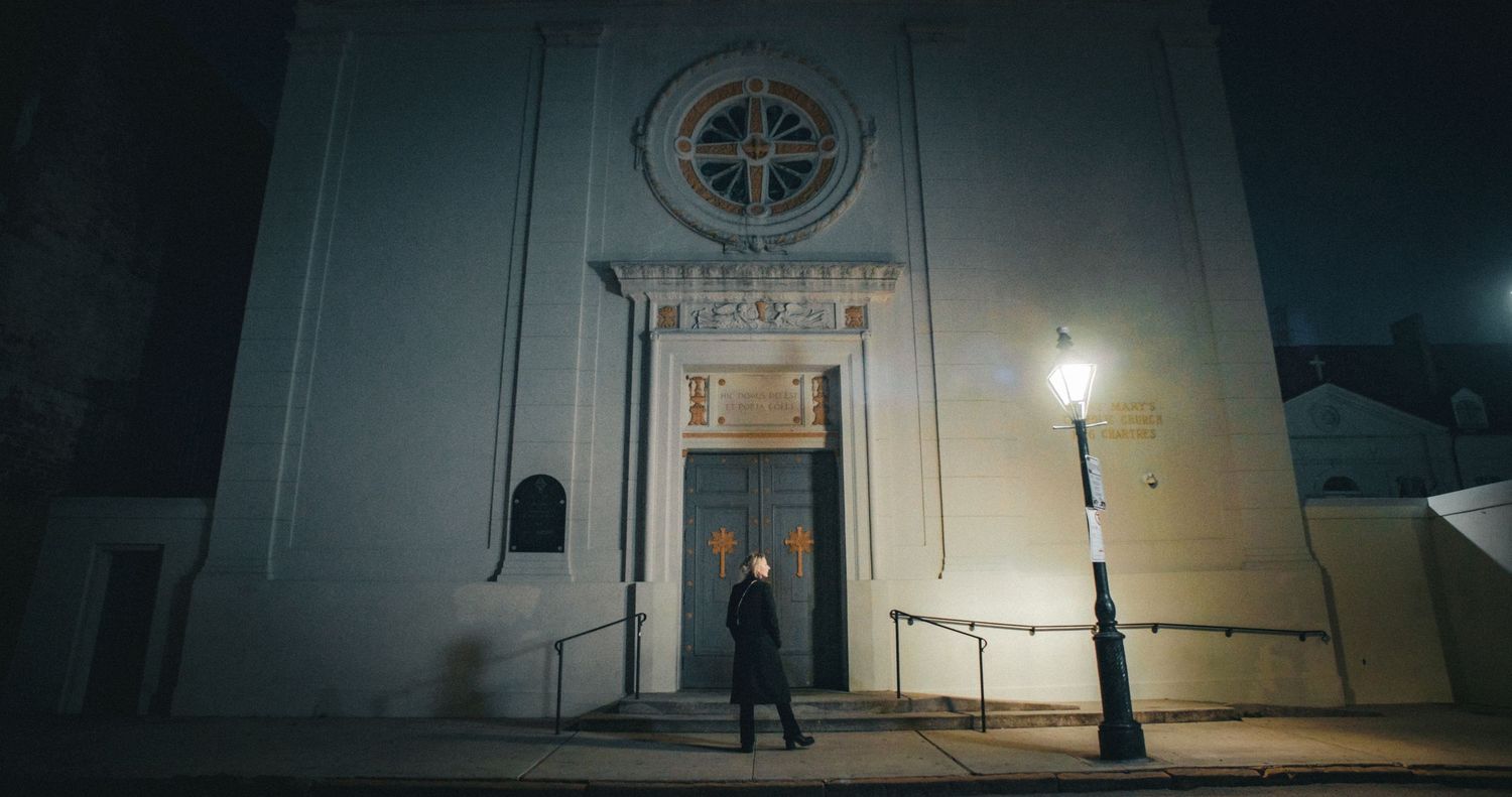Christi Lukasiak stands at entrance of an old church at night illuminated by single streetlamp. The tall facade grand cinematic atmosphere.