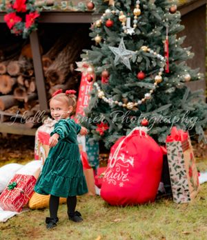 Small child in green winter coat explores decorated Christmas tree and red gift bags in rustic holiday setting.