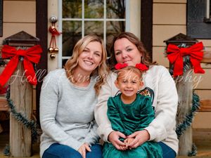 Family sits together on wooden porch steps decorated with holiday garland and red bows for Christmas.