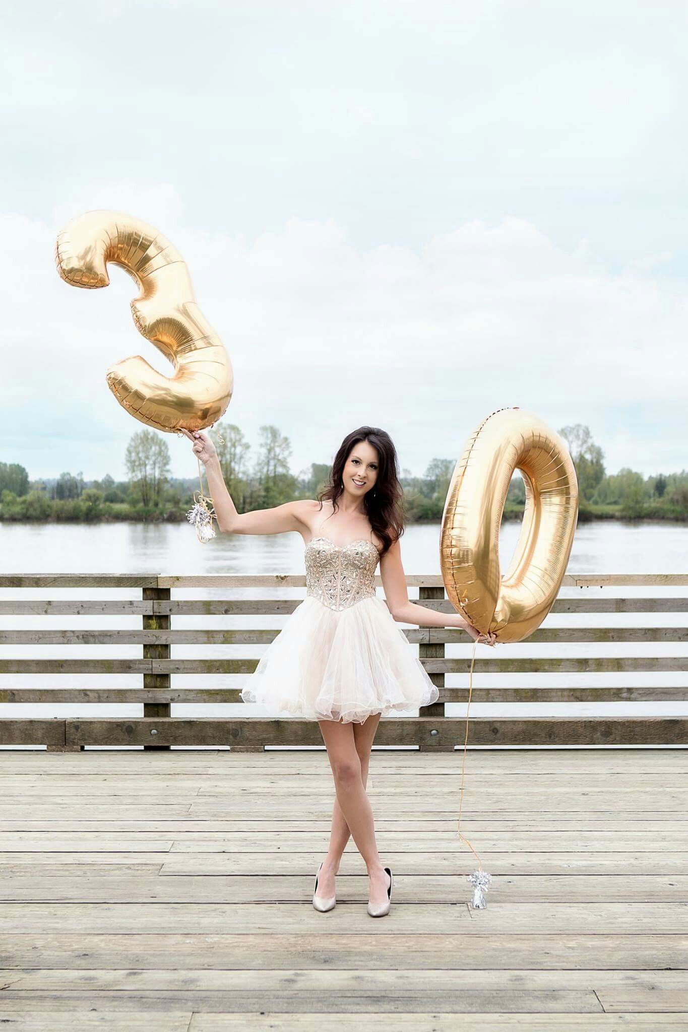 Person in white dress posing with gold number balloons on a wooden dock.