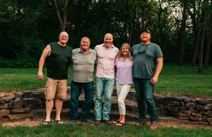 Group of people sitting together on stone wall in wooded outdoor setting.
