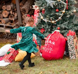 Small child in green winter coat explores decorated Christmas tree and red gift bags in rustic holiday setting.