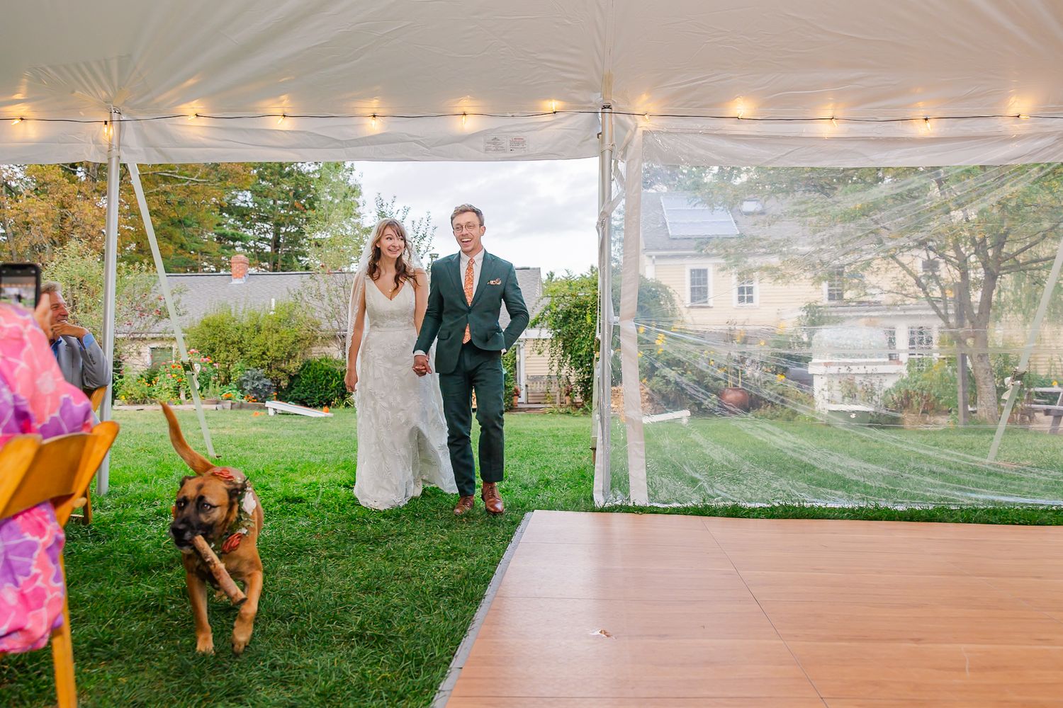 Wedding guests dance and celebrate under a white tent with string lights during an outdoor evening reception.