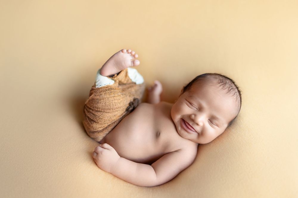 Sleeping newborn baby wrapped in beige knit blanket smiles while laying on soft cream backdrop during professional photoshoot.