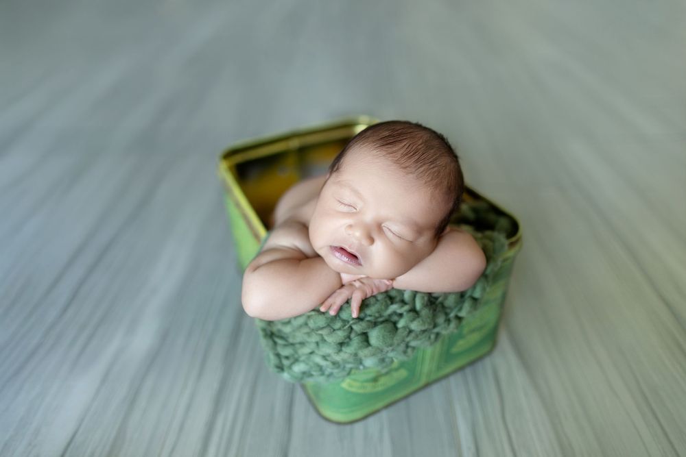 A sleeping newborn baby nestled in a green container on a light wooden floor.