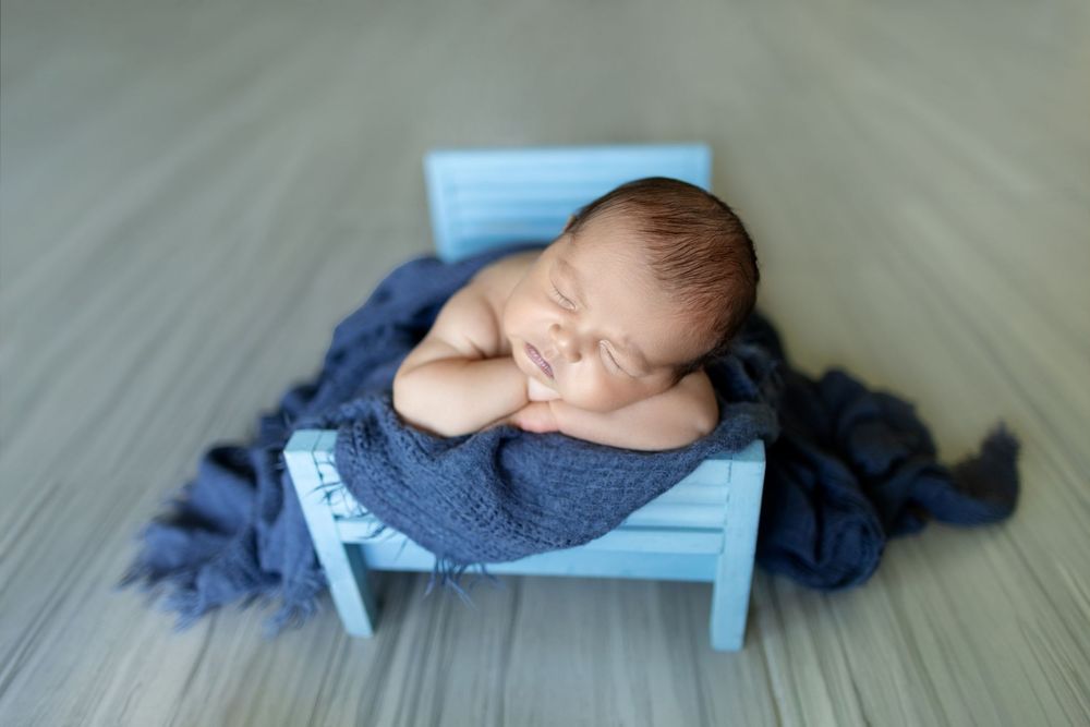 A sleeping newborn baby wearing a blue knit outfit lies curled up on a wooden floor.