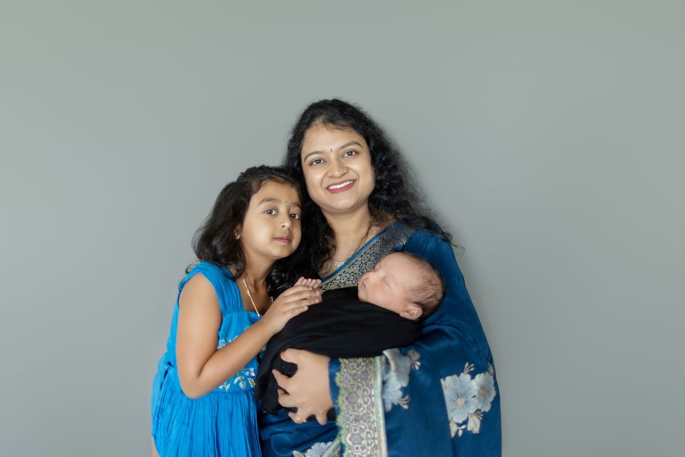 A tender family moment against a gray backdrop as a parent holds a sleeping baby while an older sibling in blue looks on.