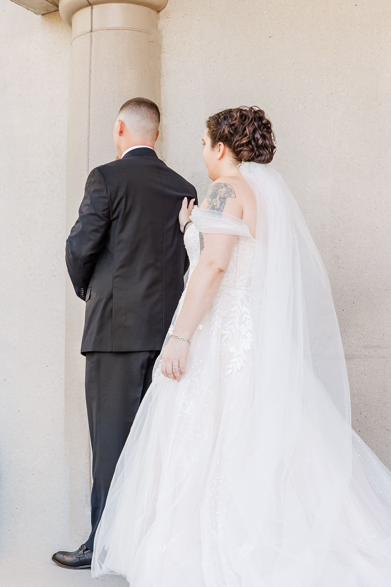 A wedding couple walks down a light-colored corridor in formal attire with a flowing white veil trailing behind.